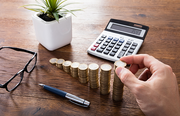 Close-up Of A Person Hand Calculating On Wooden Desk In Front Of Stacked Coins